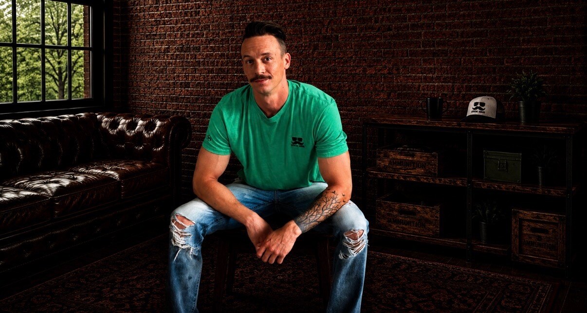 Stache Squad founder in a green Stache tee, seated in a brick-walled room beneath a hanging lamp